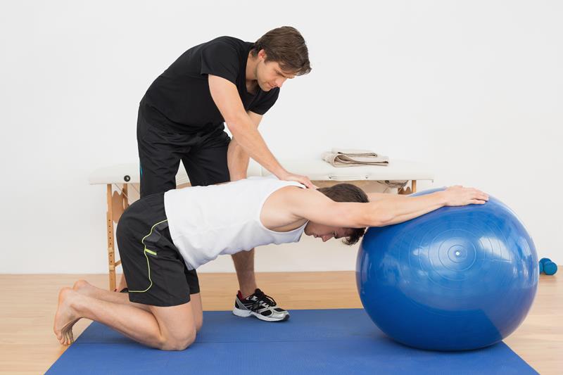 a trainer helps teach physical therapy to a patient on an exercise ball
